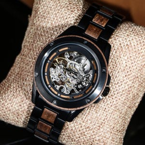 May include: A black wristwatch with a skeleton dial showing the internal mechanisms. The watch has wooden features on the bezel and strap links. It is displayed on a tan fabric surface.