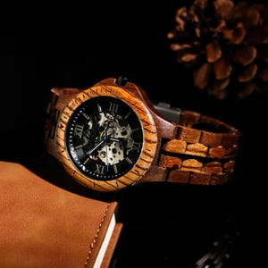 May include: A wooden wristwatch with a dark face and visible internal mechanisms. The watch has a brown wooden strap and a wooden bezel. The watch is displayed on a dark surface, with a brown leather notebook in the background.