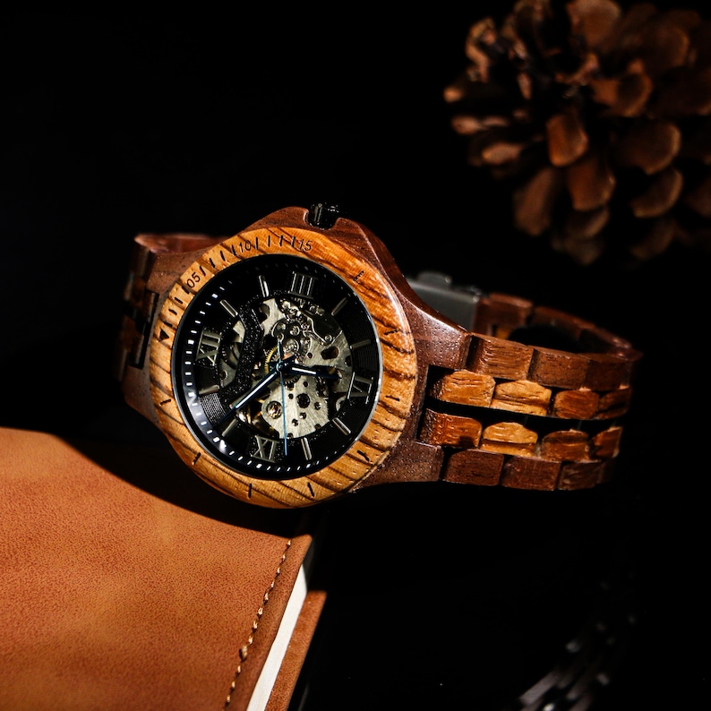 May include: A wooden wristwatch with a dark face and visible internal mechanisms. The watch has a brown wooden strap and a wooden bezel. The watch is displayed on a dark surface, with a brown leather notebook in the background.
