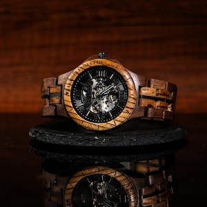 May include: A wooden wristwatch with a dark face and visible internal mechanisms. The watch has a brown wooden band and a dark bezel. The watch is displayed on a dark surface, with its reflection visible below.