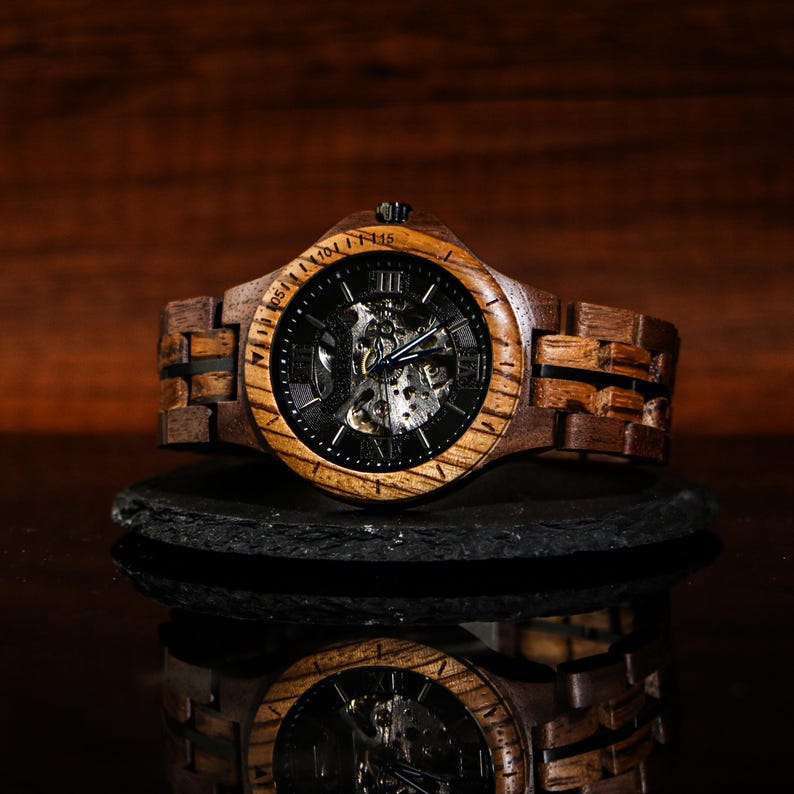 May include: A wooden wristwatch with a dark face and visible internal mechanisms. The watch has a brown wooden band and a dark bezel. The watch is displayed on a dark surface, with its reflection visible below.