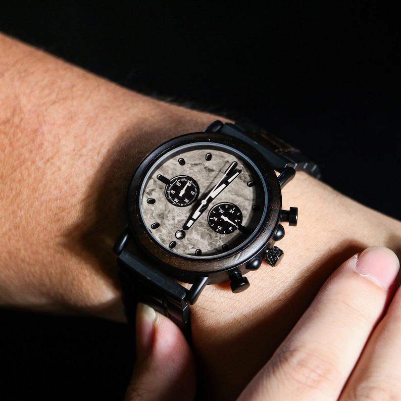 Puede incluir: Un reloj de pulsera de madera oscura con esfera veteada y detalles en negro. El reloj tiene dos esferas m&aacute;s peque&ntilde;as y funci&oacute;n de cron&oacute;metro. El reloj tiene una correa negra.