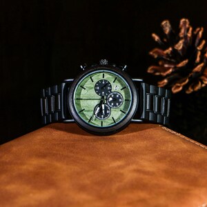 Puede incluir: Un reloj de pulsera de madera con esfera verde y detalles en negro. El reloj tiene tres subesferas y una correa de metal negro. El reloj se muestra sobre una superficie de cuero marr&oacute;n.
