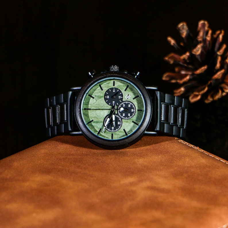Puede incluir: Un reloj de pulsera de madera con esfera verde y detalles en negro. El reloj tiene tres subesferas y una correa de metal negro. El reloj se muestra sobre una superficie de cuero marr&oacute;n.