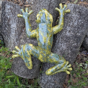 May include: A ceramic frog sculpture with a yellow and green mottled glaze. The frog is posed with its arms and legs outstretched, as if climbing. The sculpture is placed on a weathered tree trunk, with green foliage in the background.