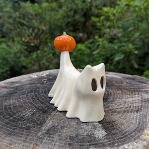 May include: A glossy off-white ceramic figurine shaped like a ghost with two oval eye cutouts. The ghost has a tall, pointed head topped with a small, bright orange pumpkin featuring a tiny green stem, creating a smooth, reflective surface finish.