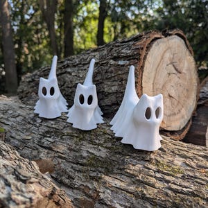 May include: Three white ceramic ghost figurines with oval eye cutouts and flowing robes are arranged on a textured log. The ghosts have a whimsical, cartoonish design, perfect for Halloween or spooky decor.