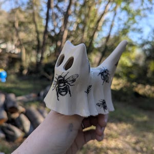 Puede incluir: Figura de gato de cerámica de color crema con ilustraciones de abejas negras. La figura tiene dos agujeros para los ojos y una cola larga y puntiaguda. El gato se sostiene en una mano sobre un fondo borroso.