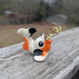 Può includere: Una statuetta di pesce koi in ceramica, dipinta in bianco, arancione e nero, con fiori secchi. Il pesce è posizionato su una superficie di legno stagionato, con uno sfondo esterno sfocato.