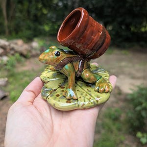 May include: A ceramic frog figurine with a brown barrel on its back, perched on a green lily pad. The frog is green and brown with black eyes. The barrel is a reddish-brown color. The figurine is held in a hand.