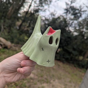 May include: A light green ceramic ghost figurine with a red mouth and black eyes. The ghost has a pointed top and a cross and an X on the front. The figurine is held in a hand against a blurred outdoor background.