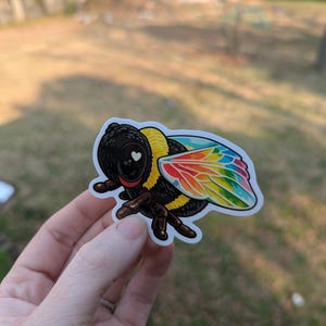 May include: A sticker of a cartoon bee with a black body, yellow stripes, and rainbow-colored wings. The bee has a heart-shaped detail on its face. The sticker is held up against a blurred outdoor background.