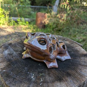 May include: A brown ceramic frog figurine with a glossy finish and multiple circular cutouts across its back. The frog has large, dark, smooth eyes and a wide mouth, with its legs splayed outward in a resting position on a flat surface.