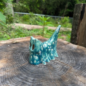 May include: A turquoise ceramic ghost figurine with a textured surface and white accents. The ghost has two eye holes and a pointed top. The figurine is displayed on a wooden surface with a natural background.