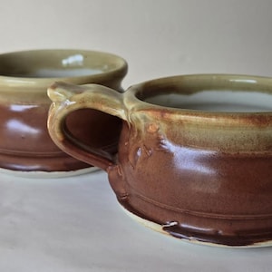 May include: Two handmade ceramic mugs with handles. The mugs have a brown base, a tan rim, and a white interior. The mugs are sitting on a white surface. These mugs are perfect for coffee, tea, or soup.