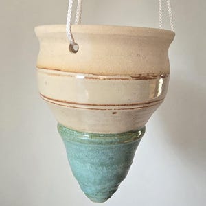 May include: A hanging ceramic planter with a cream-colored top, a light brown band, and a turquoise bottom. The planter is suspended by white rope. This handcrafted piece is ideal for displaying small plants or succulents.