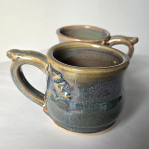May include: Two handmade ceramic mugs with a unique, textured design. The mugs feature a dark blue and brown glaze with hints of green and gold. Each mug has a curved handle and a slightly flared rim.