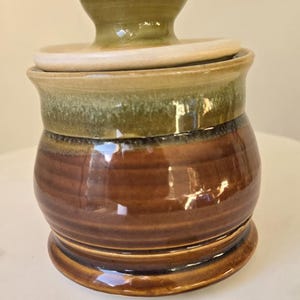 May include: A handcrafted ceramic jar with a lid. The jar features a rounded body with a brown base, a green midsection, and a tan lid and handle. The jar is approximately 4 inches tall and 4 inches wide.