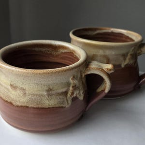 May include: Two handmade ceramic mugs. The mugs have a rounded shape with a wide opening and a handle. The exterior features a two-tone color scheme, with a reddish-brown base and a beige upper section. The interior is a dark brown color.
