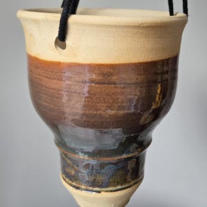 May include: A handmade ceramic hanging planter with a natural, earthy aesthetic. The planter features a cream-colored rim, transitioning to a rich brown midsection, and a dark, glazed base. Black rope suspends the planter, ready for display.