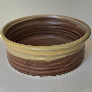 May include: A handmade ceramic bowl with a wide, shallow design. The bowl features a layered color scheme, with a light yellow rim, a band of tan, and a deep brown base. The interior is a solid brown color.