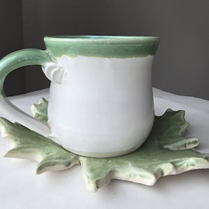 May include: A handmade ceramic mug with a white body and a green rim and handle. The mug sits on a leaf-shaped coaster, also in shades of green. The mug is designed for hot beverages.