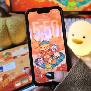 Puede incluir: Un teléfono inteligente negro muestra un fondo de pantalla con temática otoñal con personajes de dibujos animados. La pantalla muestra la hora 5:50. Una calabaza de peluche y una luz nocturna con forma de pato están en el fondo.