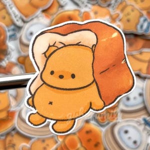 Op de afbeelding: Een verzameling stickers met een cartoon beer karakter met een brood-achtig uiterlijk. De beer is lichtoranje met een kleine buik en zwarte ogen. De stickers hebben een witte rand en zijn verspreid over de afbeelding.