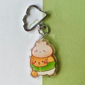 May include: A clear acrylic keychain featuring a cartoon dumpling character with a smiling face, wearing a green sweater. The keychain has a silver-colored cloud-shaped clasp and a ring. The text "gubu.family" is printed on the keychain.