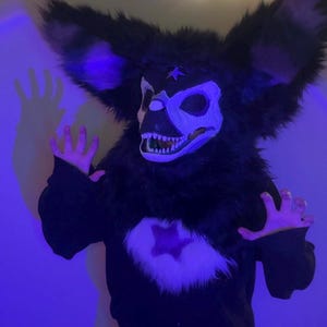 May include: A full-head creature mask featuring long black faux fur, large pointed ears, and a white skull-like face with sharp teeth. The mask includes a black star symbol on the forehead and a black sweatshirt with a white star patch on the chest.