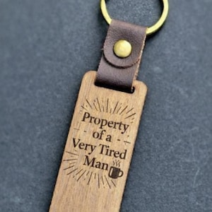 Puede incluir: Llavero de madera con un anillo dorado y una correa de cuero marrón. La etiqueta rectangular de madera está grabada con la frase "Property of a Very Tired Man" y un icono de taza de café.