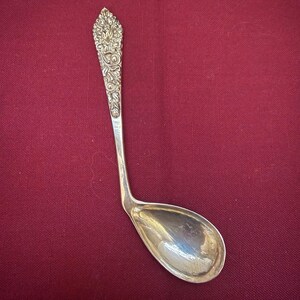 May include: A silver-toned metal spoon featuring an ornate, embossed floral pattern on the handle. The handle is long and slender, transitioning into a wide, rounded bowl that is angled sharply to the side, creating a distinct, asymmetrical shape.