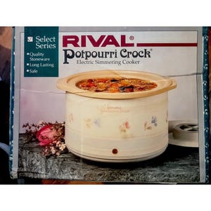 May include: A Rival Select Series Potpourri Crock electric simmering cooker in a box. The stoneware crock is cream-colored with a floral pattern, featuring a tan lid and a small power indicator light. The box displays the product name and text.
