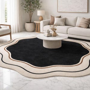 May include: A black, irregularly shaped rug with a cream border and brown and black accent lines. The rug is in a living room with a white sofa, armchair, and coffee table. The rug's unusual shape adds a modern touch to the room.