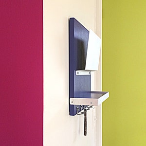 May include: A dark blue wall-mounted shelf with a white shelf and a white magazine holder. The shelf has five hooks and a small wooden object hanging from one of the hooks. The wall is painted in a bright pink and yellow color.