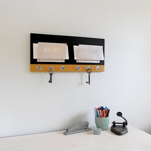 May include: A black and gold key rack with two mail slots labeled "BRIAN" and "INGLA". The rack has six hooks with two keys hanging from them.