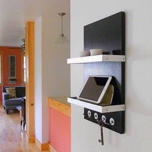 May include: A black and white wall mounted shelf with a white shelf and a black back panel. The shelf has a tablet on it and a row of hooks below. There are two small white bowls on the top shelf.