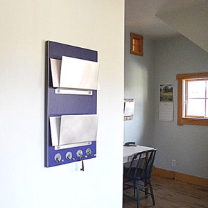 May include: A wall-mounted mail organizer with two metal mail slots and a key rack. The organizer is painted a deep blue color and has a silver metal finish. The key rack has four hooks and a single key is hanging on one of the hooks.