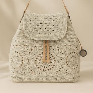May include: A cream-coloured crochet backpack with a flap closure and drawstring. The bag features a detailed circular pattern on the front and sides, with tan leather straps and a small circular charm.
