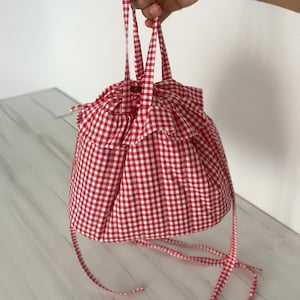 May include: A red and white gingham drawstring bag with a ruffled top. The bag is held by its straps, showcasing its round shape and the pattern of the fabric. The bag's straps are also made of the same gingham fabric.
