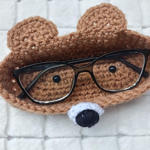 May include: A handmade, brown crocheted glasses holder shaped like a bear's face, complete with ears, a white and black nose, and black-framed eyeglasses inside. The holder sits on a white, textured surface.