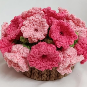 May include: A handmade crochet floral arrangement in a basket. The flowers are various shades of pink, with green leaves, and the basket is brown. The flowers are densely packed, creating a full, textured appearance. The craftwork is detailed and intricate.