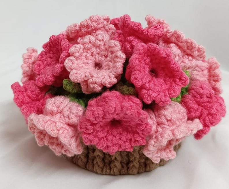 May include: A handmade crochet floral arrangement in a basket. The flowers are various shades of pink, with green leaves, and the basket is brown. The flowers are densely packed, creating a full, textured appearance. The craftwork is detailed and intricate.