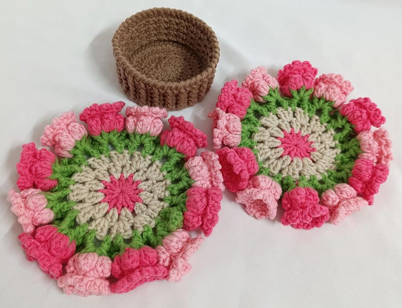 May include: Two handmade crochet coasters with a floral design in pink, green, and beige, alongside a small brown crochet basket. The coasters feature a central pink flower surrounded by green and beige petals, with pink flower accents.