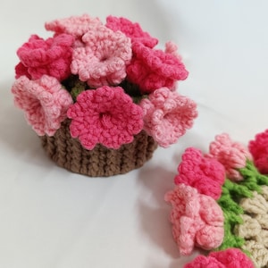May include: A handmade crochet flower arrangement in a brown basket. The flowers are in shades of pink and red, with green leaves. The arrangement is on a white surface. The flowers are made of yarn.