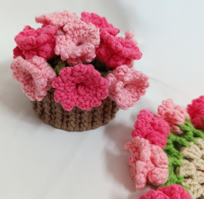 May include: A handmade crochet flower arrangement in a brown basket. The flowers are in shades of pink and red, with green leaves. The arrangement is on a white surface. The flowers are made of yarn.