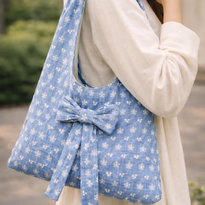May include: A blue tote bag with a white floral pattern and a large bow. The bag has a knotted strap and is made of a soft fabric. The bag is a shoulder bag.