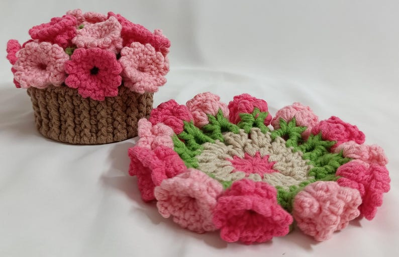 May include: Handmade crochet flower basket and coaster set. The basket is brown with pink and light pink flowers. The coaster has a floral design with pink, green, and beige colors. The set is made of yarn.