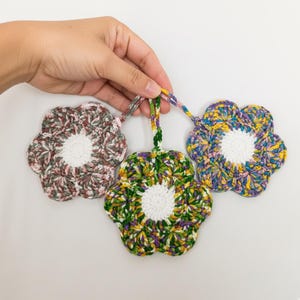 May include: Three handmade crochet flower ornaments in shades of green, purple, yellow, and brown. Each flower has a white center and a loop for hanging. The ornaments are held by a hand against a white background.