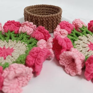 May include: A handmade crochet set featuring a brown basket and floral coasters. The coasters have a pink, green, and cream color scheme, with a flower design. The basket is a solid brown color.